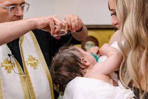 A priest performing a baptism ceremony on an infant held by a woman in a white dress.
