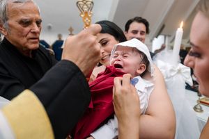 A crying newborn being baptized by an elderly priest while being held by a woman.