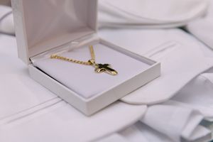 A gold cross necklace in a white jewelry box placed on white baptism garments.