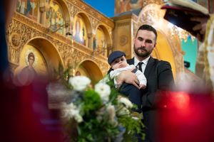 A bearded man in a suit holds an infant inside a ornate Orthodox church during a baptism.
