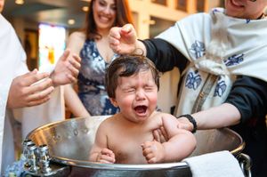 Baby crying during Orthodox baptism immersion in a silver baptismal bowl held by a priest.