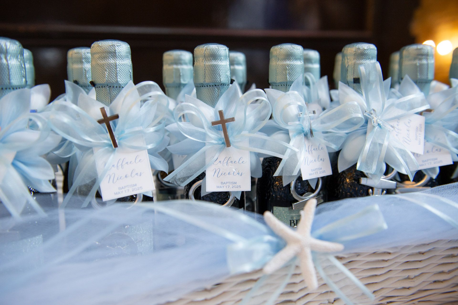 Blue ribbon-tied olive oil baptism favors with cross tags and starfish decoration in a wicker basket.