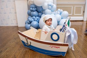Adorable baby in white baptism outfit sitting in a decorative wooden toy boat with blue balloon backdrop.
