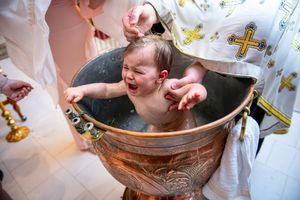 A crying baby being baptized by immersion in a copper font by an Orthodox Christian priest.