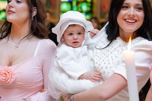 A baby in a white christening outfit and bonnet held by a smiling woman during a baptism ceremony.