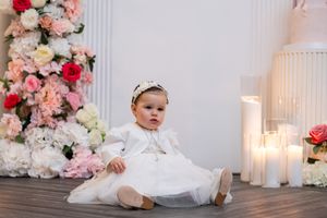 A baby in a white christening dress sits on the floor surrounded by flowers and candles for a portrait.