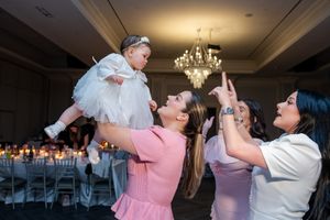 Two women in pink and white dresses lift a baby in white at an elegant baptism reception.