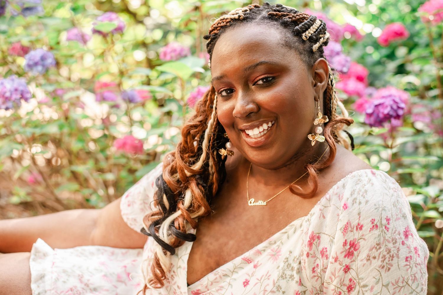 Smiling young Black woman with long red braids wearing a blue top and gold necklace, leaning against a wall outdoors.