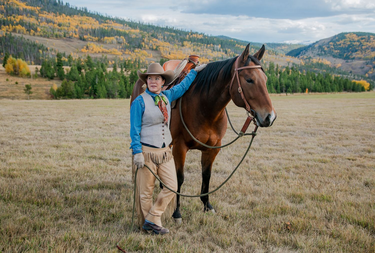 A young cowboy stands beside a bay horse in an autumn mountain meadow, wearing a hat and western attire.