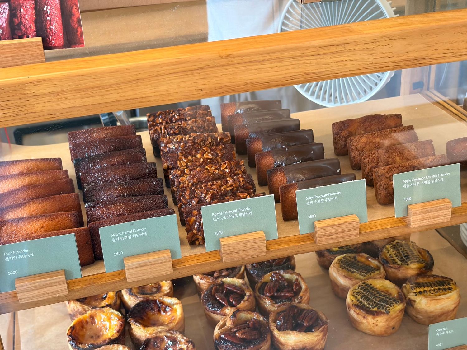 Bakery display case with chocolate brownies, pecan bars, and golden egg tarts arranged on wooden shelves with label cards.