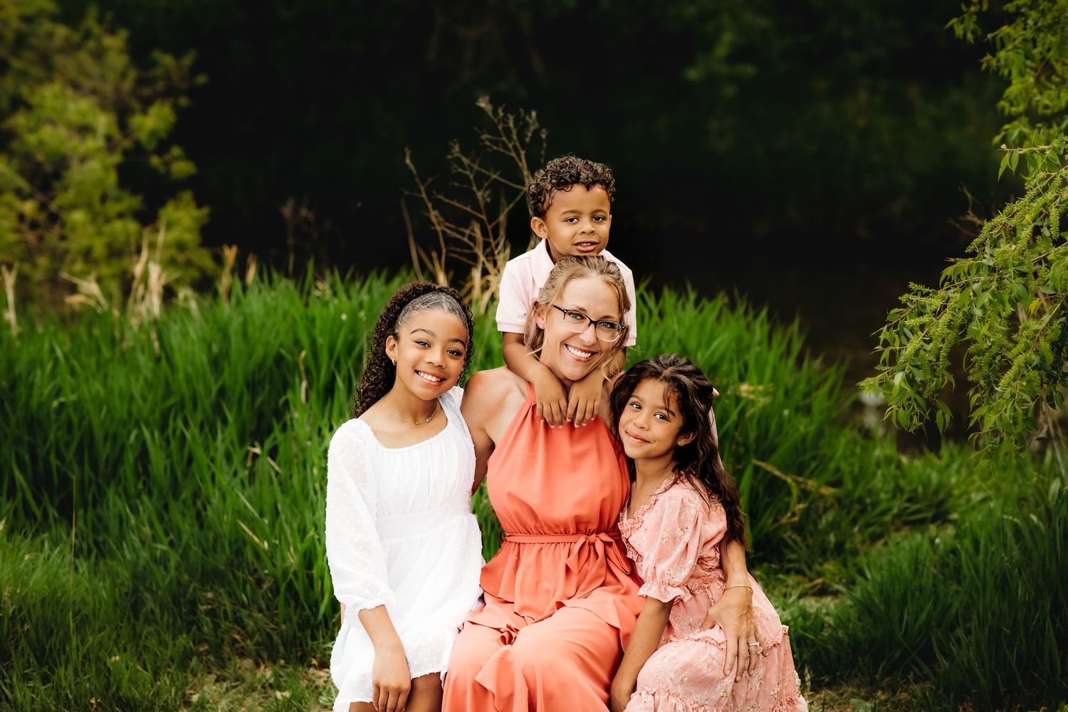 A group poses together in a lush green outdoor setting during golden hour wearing coral and white outfits.