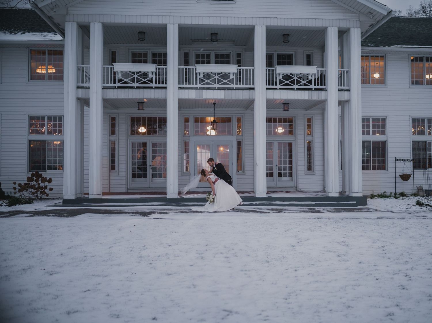 A romantic winter wedding scene in front of a grand white colonial mansion with snow-covered grounds and glowing windows.