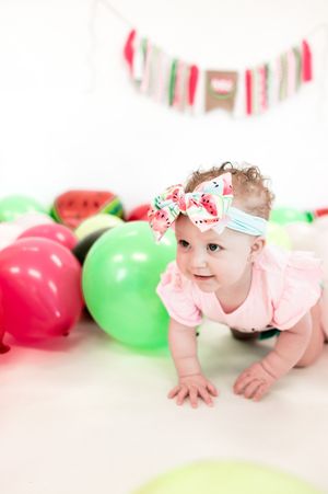 A baby in a pink outfit crawls on a white floor with red and green balloons in the background during a party setup.