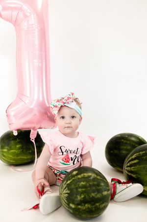 First birthday photo setup with pink balloon number one, watermelons, and baby wearing floral headband and pink outfit.