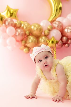 Baby in yellow fairy wings and tiara poses against pink background with gold and white balloon decorations.