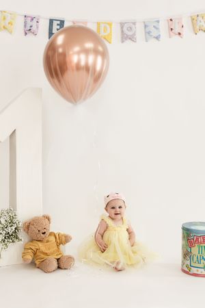 A baby in yellow dress and teddy bear sit on white floor with pink balloons and birthday decorations above.