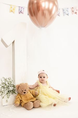 Baby in yellow dress lies on white floor with teddy bear and pink balloons during first birthday photo session.