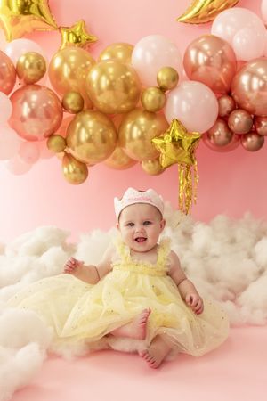 Baby in yellow tulle dress lying on cloud-like setting with pink and gold balloon arch decorations for milestone photo shoot.