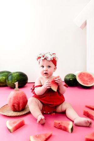Baby in red outfit poses with fresh watermelon slices on pink background in summer-themed photo series.