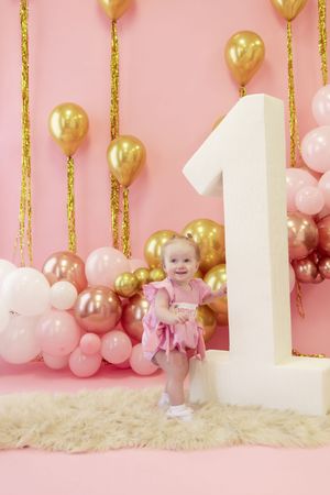 Pink and gold first birthday party setup with white number one decoration, balloons and glittery streamers against pink backdrop.