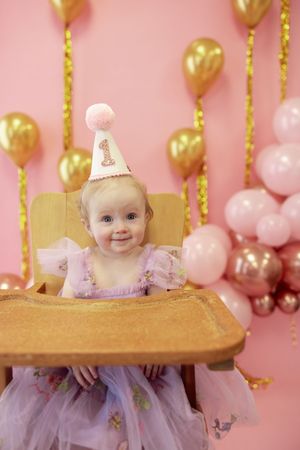 First birthday celebration with pink and gold balloons featuring a baby in a tulle dress sitting in wooden high chair.