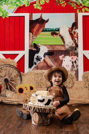 Child in overalls and cowboy hat sits by farm display with rustic wood decor and red barn background.