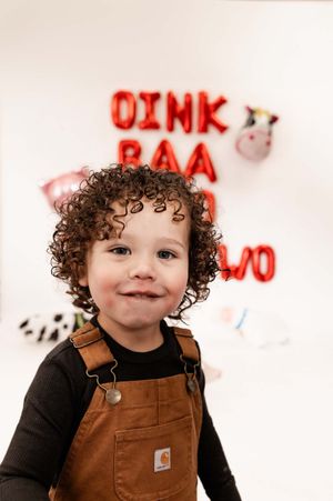 A young child in brown Carhartt overalls and black shirt smiles against a white wall with red graffiti.