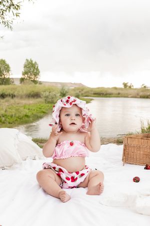 Baby in strawberry print outfit sitting on white blanket with picnic basket outdoors.