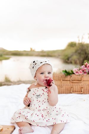 Sweet summer picnic scene with a baby in a cherry-print dress enjoying fruit on a white blanket beside a picnic basket.