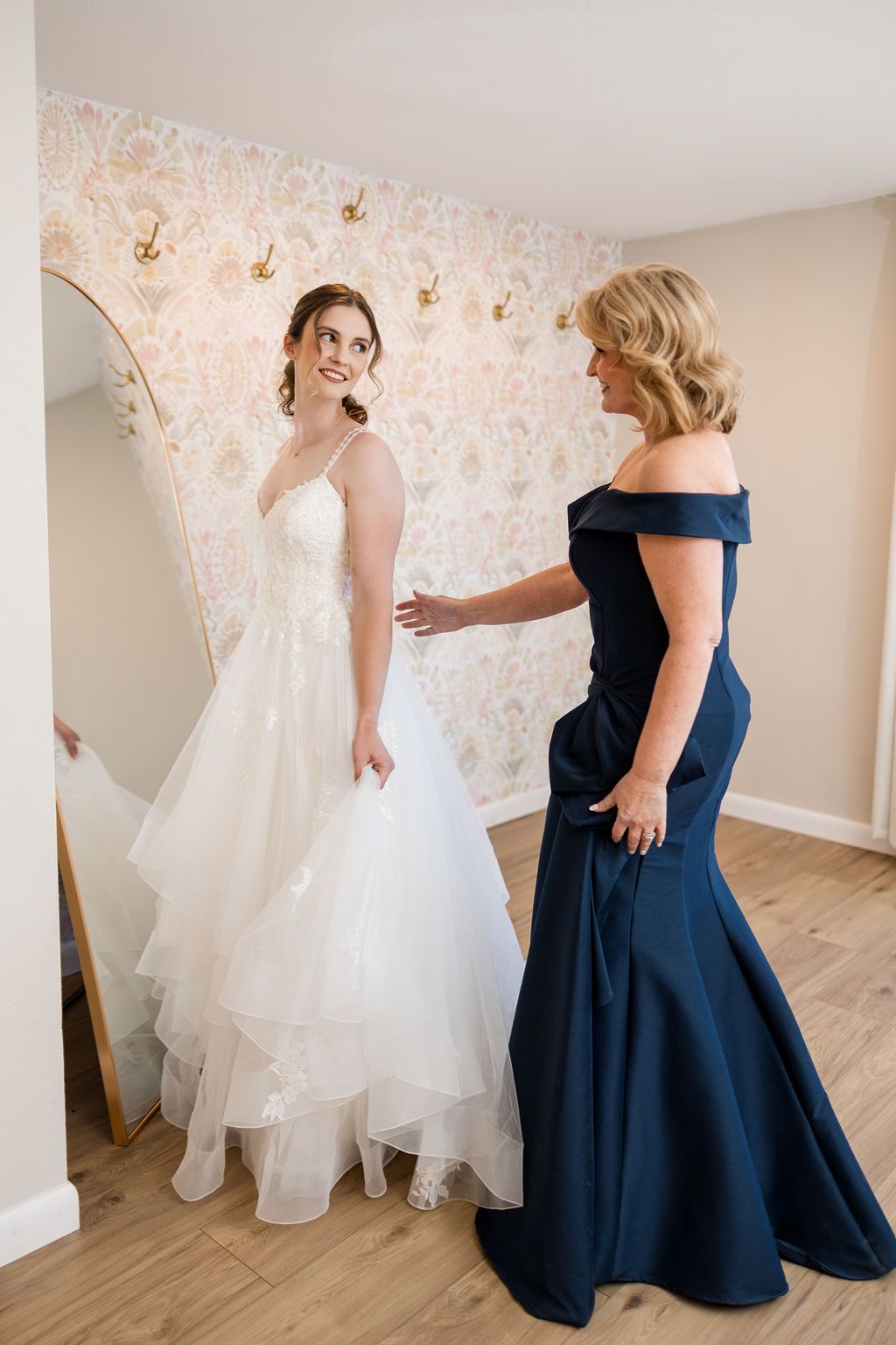 A flowing white wedding dress with intricate lace details and long train displayed in a sunlit room with hardwood floors.