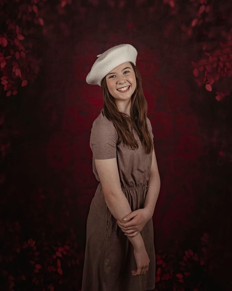 A person in a brown dress poses against a dark red backdrop with dramatic lighting and floral elements in the background.