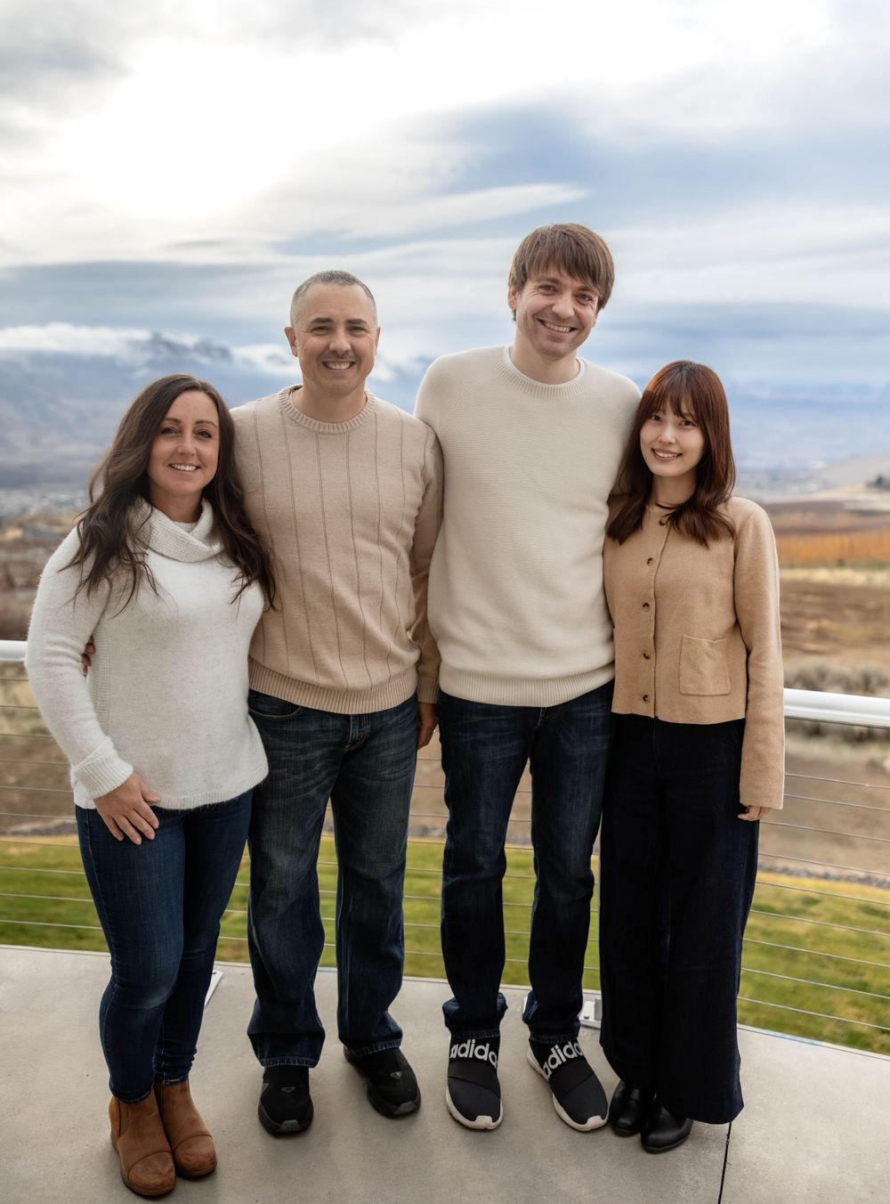 A family wearing coordinated neutral sweaters poses together against a mountain landscape backdrop.