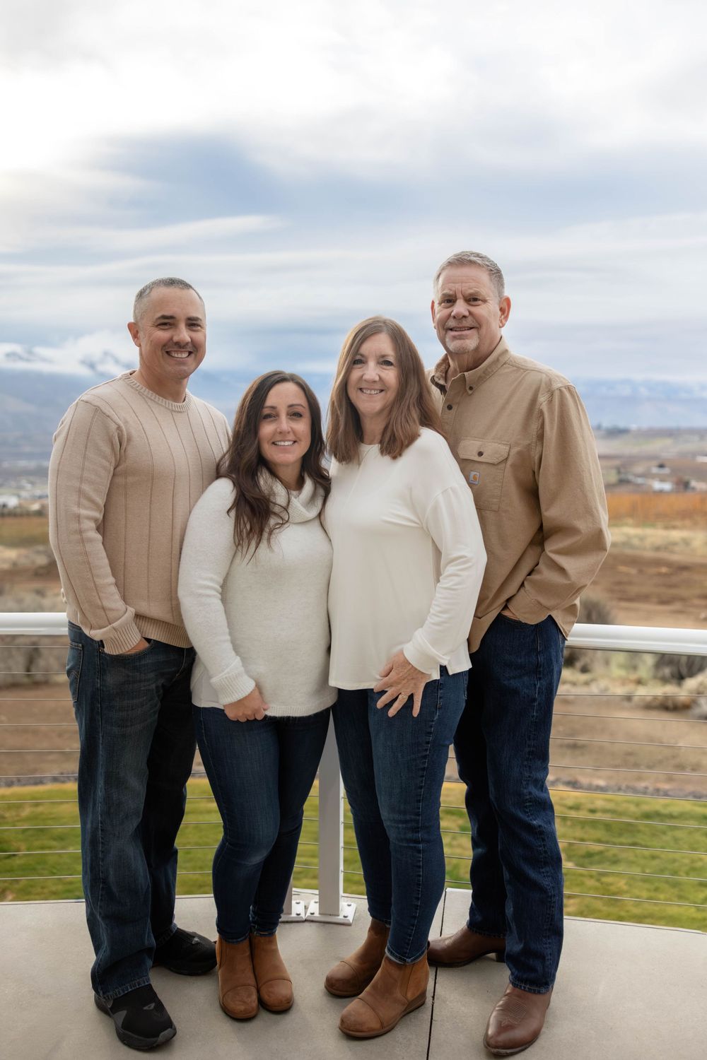 Family group wearing matching cream sweaters and jeans stands together on an outdoor patio.