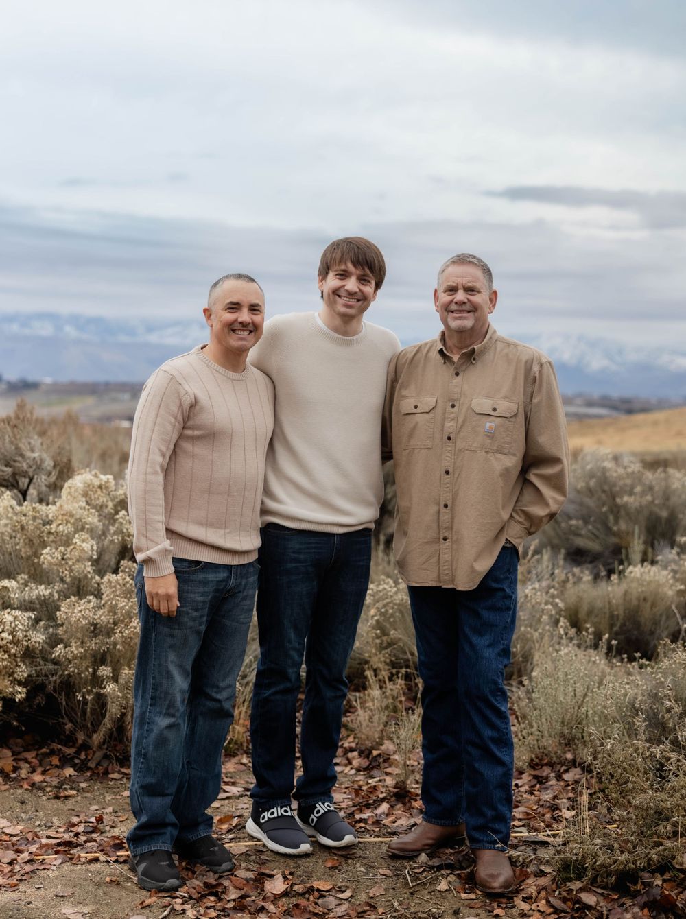 Three people in coordinating beige sweaters pose together in a desert landscape setting.