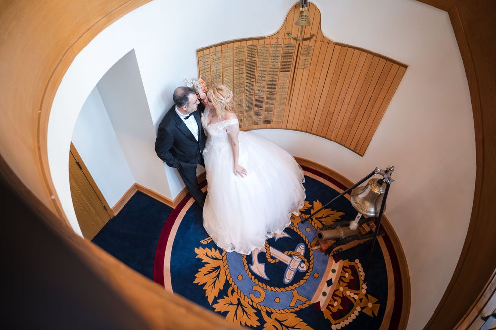 A couple in wedding attire stands on a grand spiral staircase with ornate blue and gold carpeting viewed from above.