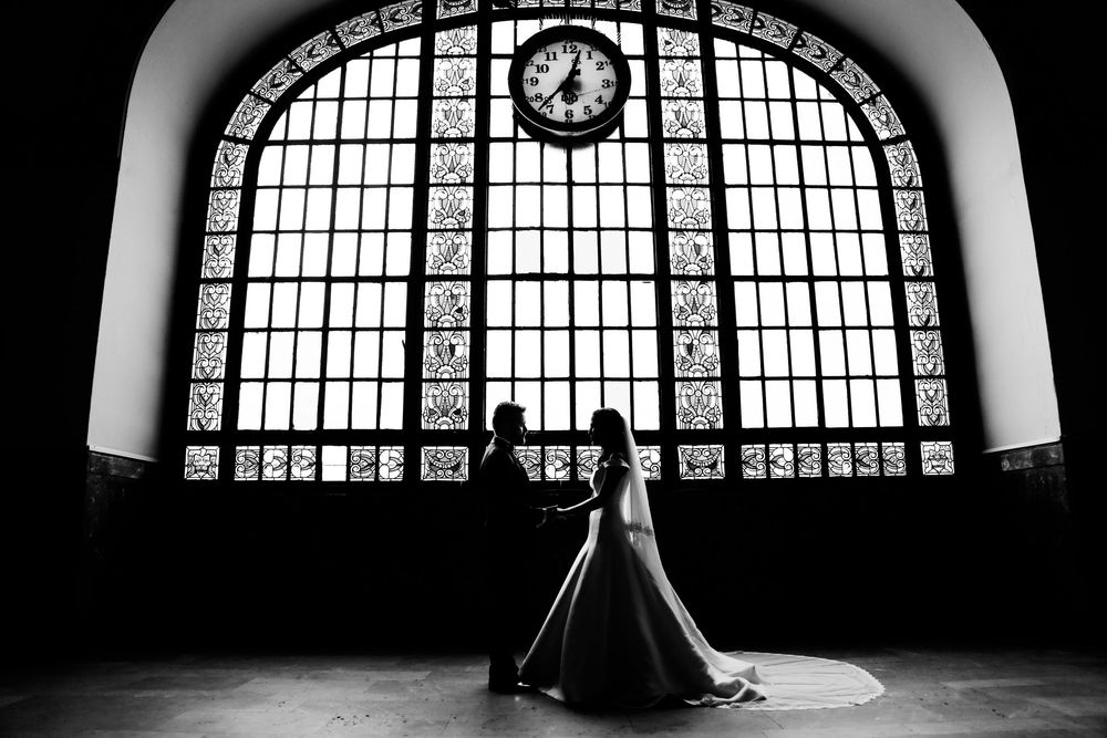 Dramatic silhouette of a couple dancing in front of a grand arched window with a clock in a vintage train station.