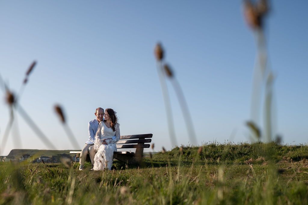 Just married couple sit on a bench overlooking Bude beach. Photograph is from a distance looking through grass