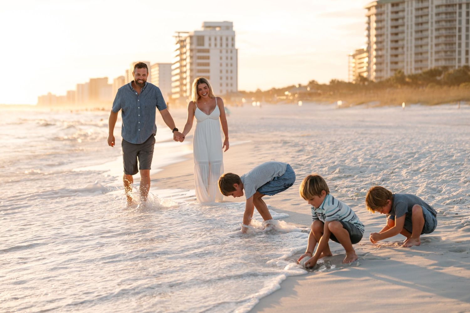 A couple walks hand in hand along a pristine white sandy beach at sunset with high-rise buildings in the background.