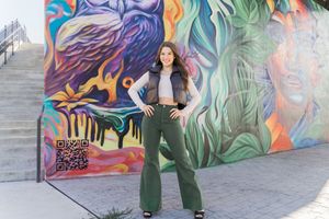 Someone poses in front of a vibrant street art mural featuring colorful abstract designs and swirling patterns.