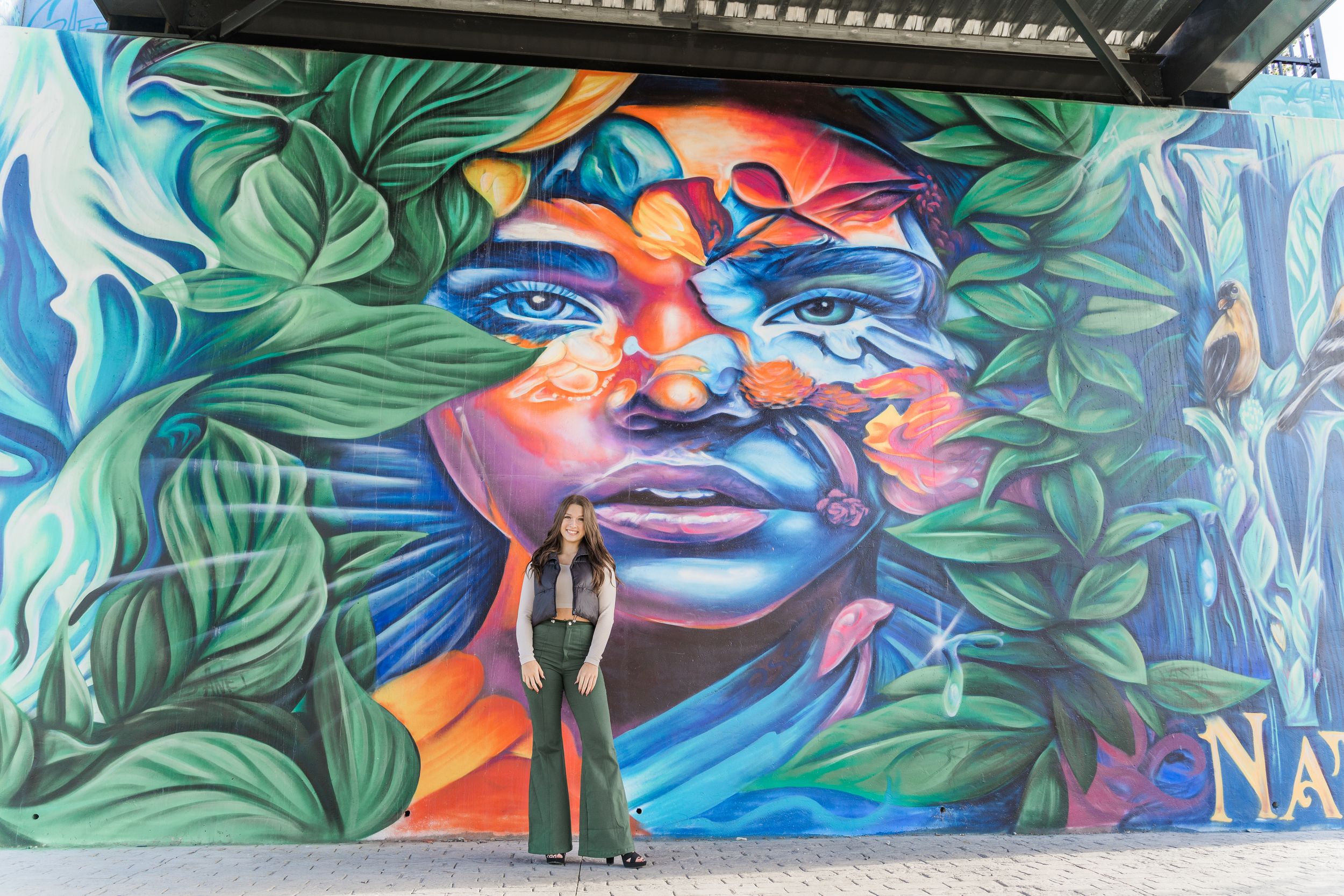 Large-scale street art mural displays vibrant faces and organic forms in blue, green, and orange tones.