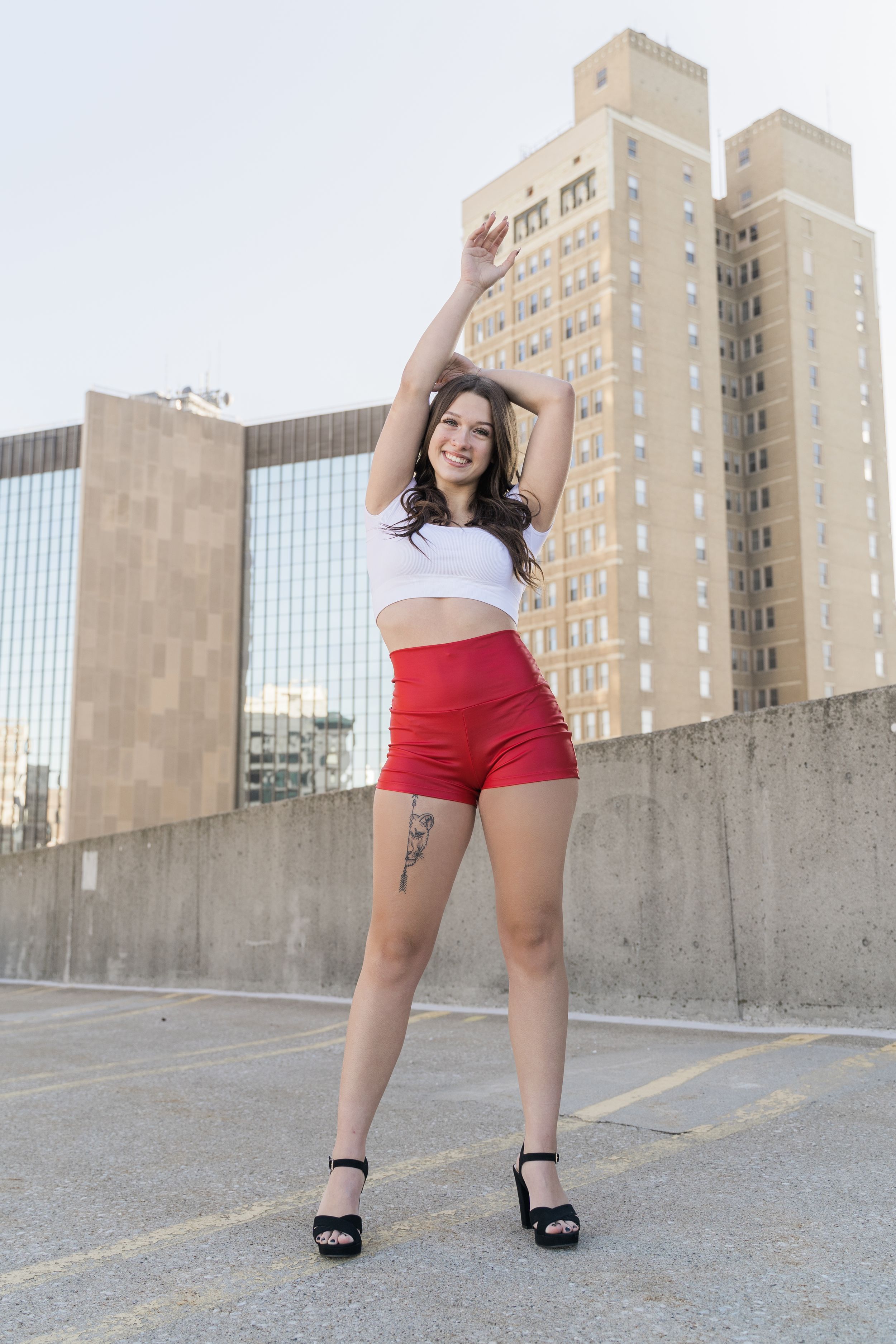 Woman in red shorts and black top poses playfully on urban rooftop with city buildings in background.