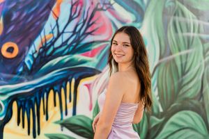 Woman in lavender dress poses in front of vibrant graffiti wall with tropical leaf designs.