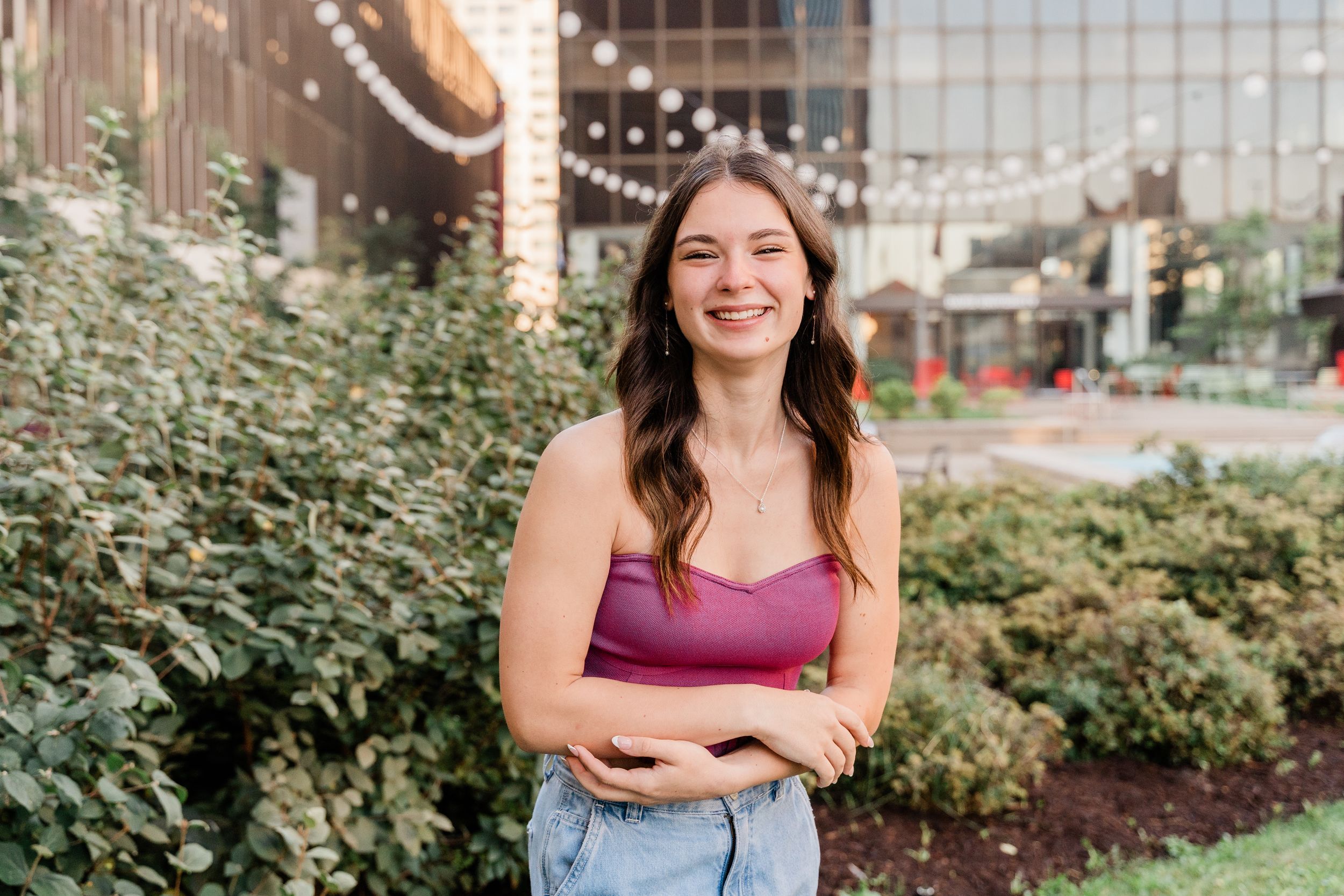 A person in a burgundy top smiles while standing outdoors near green bushes and a glass building structure.
