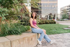 A person in casual attire sits on a stone ledge in an urban garden setting with tall buildings in the background.