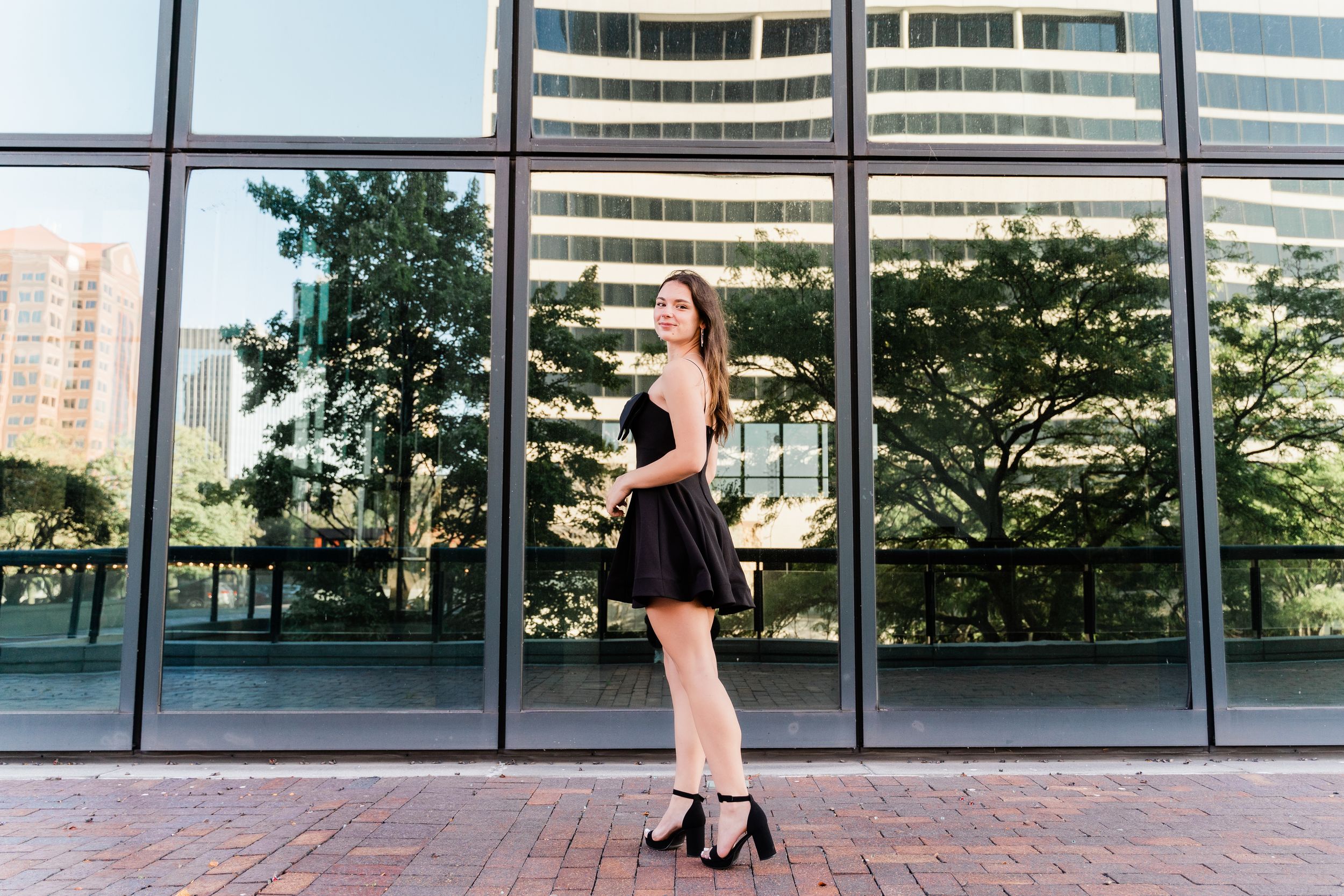 A woman in a black dress and heels walks past a modern glass office building exterior on a sunny day.