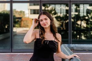 Woman in black dress poses confidently outside modern glass building during sunset photoshoot.