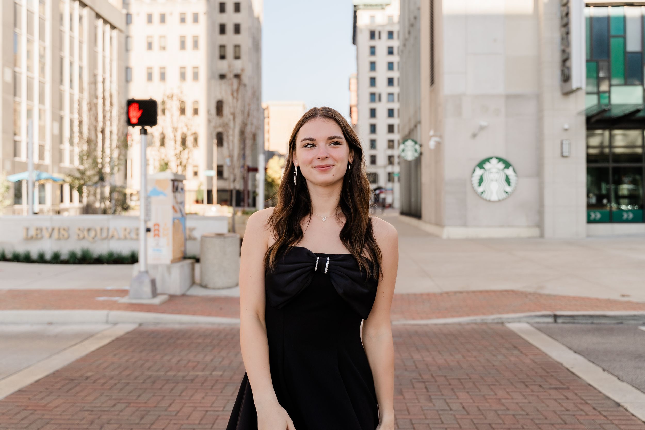 A person in a black sleeveless dress stands confidently against an urban backdrop with tall buildings.