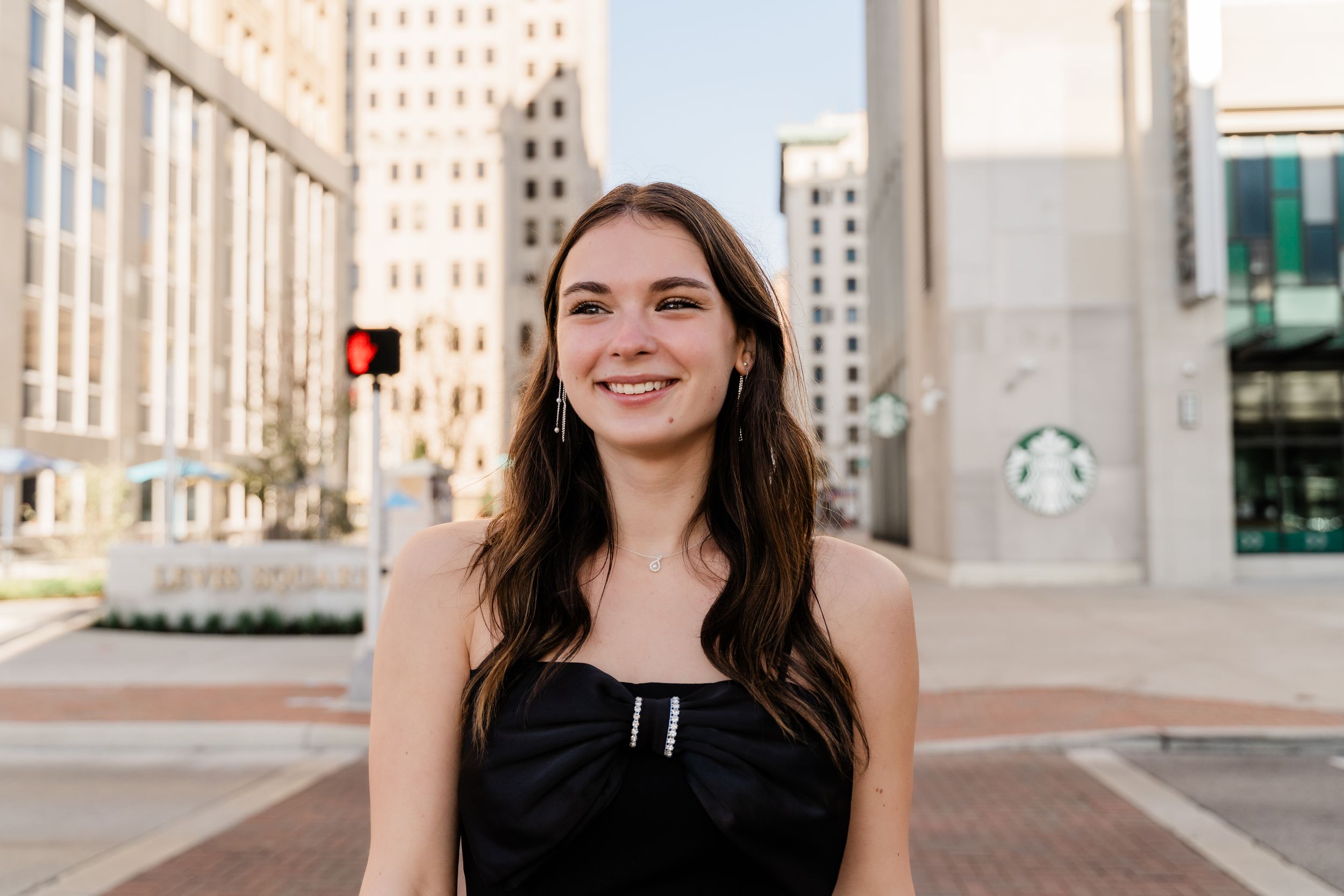 A person in a black dress smiles while standing in front of a modern urban building exterior.