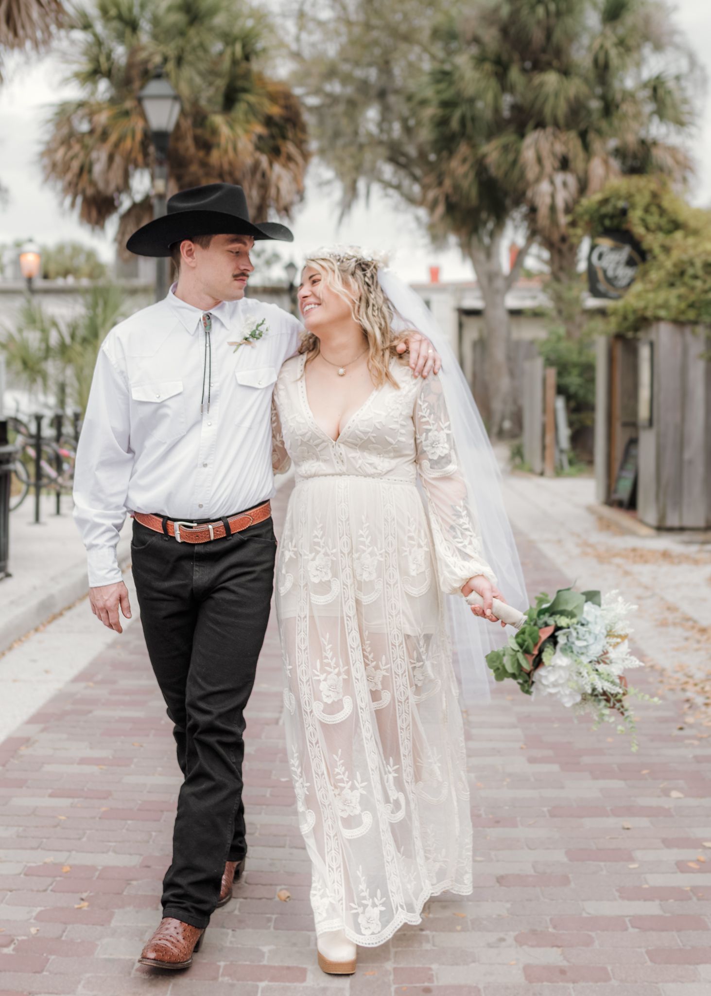 Couple in western attire walking hand in hand along a palm-lined brick pathway in a romantic outdoor setting.