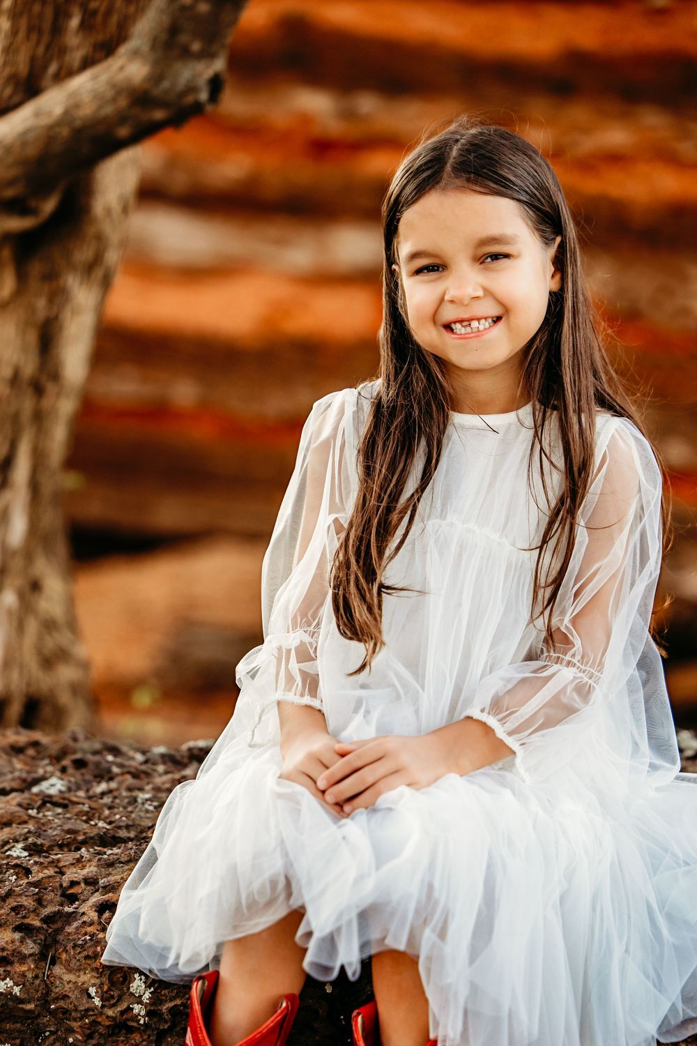 A child in a white flowy dress sits outdoors during autumn with warm orange tones in the background.